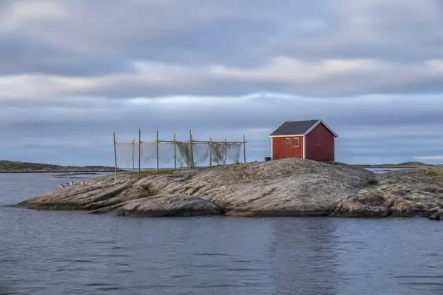 A small red fishing hut and drying nets on a bare rocky skerry in a calm grey-blue sea.