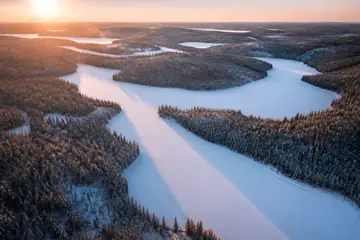 Snow-covered frozen lakes wind between dark snowy forest toward a low winter sun on the horizon.