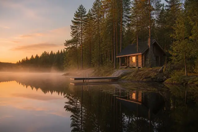 A small log sauna with a stone chimney stands by a misty lake with a narrow jetty and ladder.