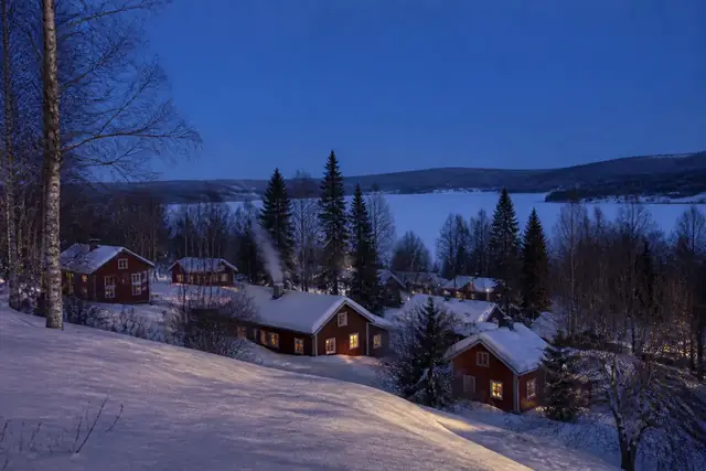 Red-ochre wooden houses with lit windows and smoking chimneys on a snowy slope above a frozen lake.